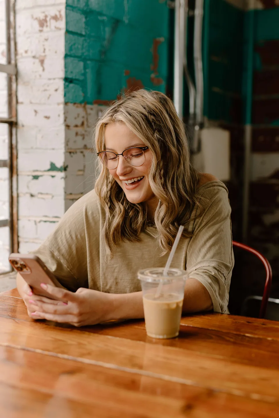 A girl laughing while holding her phone and sitting on cafe table.