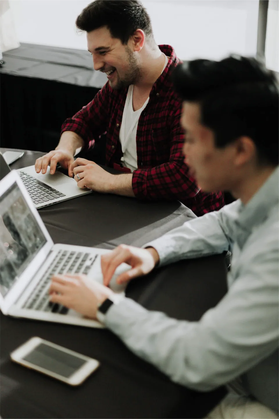 Two men working on a table with laptops in front of them.